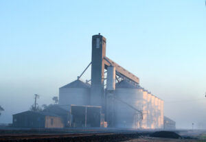 A large grain storage facility with silos and an elevator tower, surrounded by morning mist along a railway line in the Corn Belt.