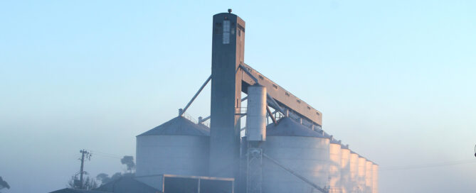 A large grain storage facility with silos and an elevator tower, surrounded by morning mist along a railway line in the Corn Belt.
