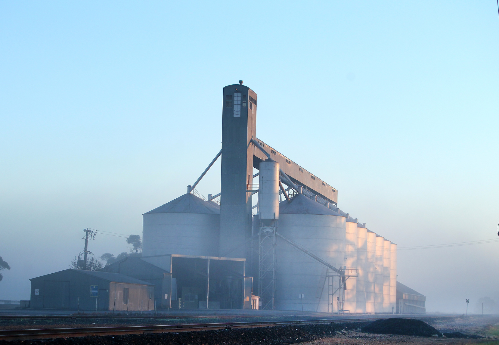 A large grain storage facility with silos and an elevator tower, surrounded by morning mist along a railway line in the Corn Belt.